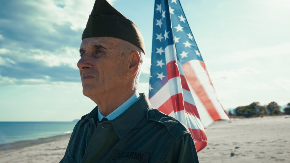 Older man in a military-style uniform stands on a beach with a large American flag behind him.