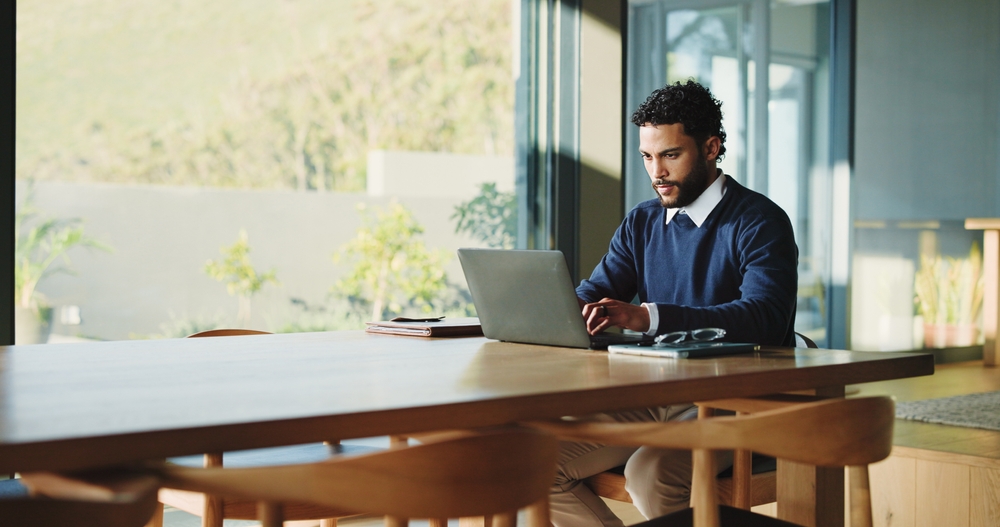 Man working on a laptop at a long wooden table beside large sunlit windows.