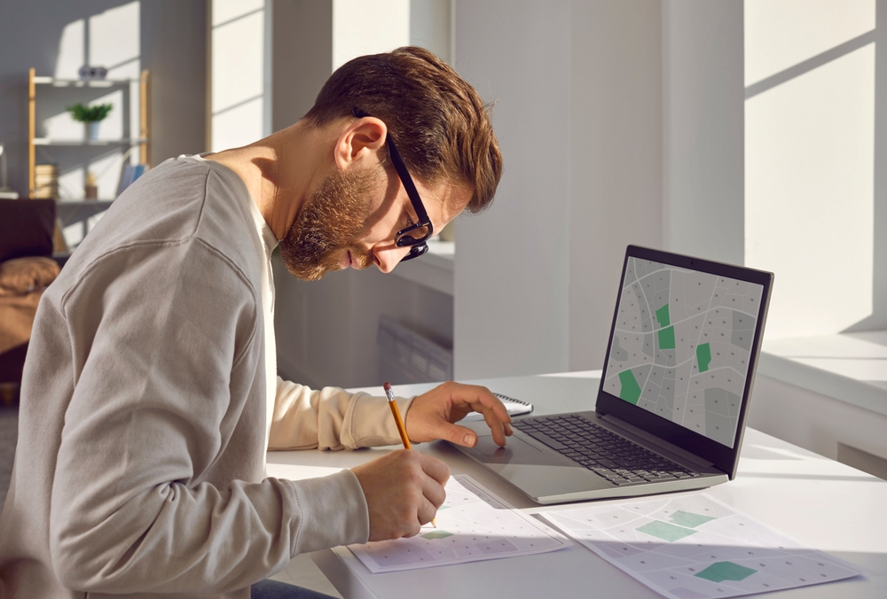 Man wearing glasses writes at a desk beside a laptop showing a map-like screen.