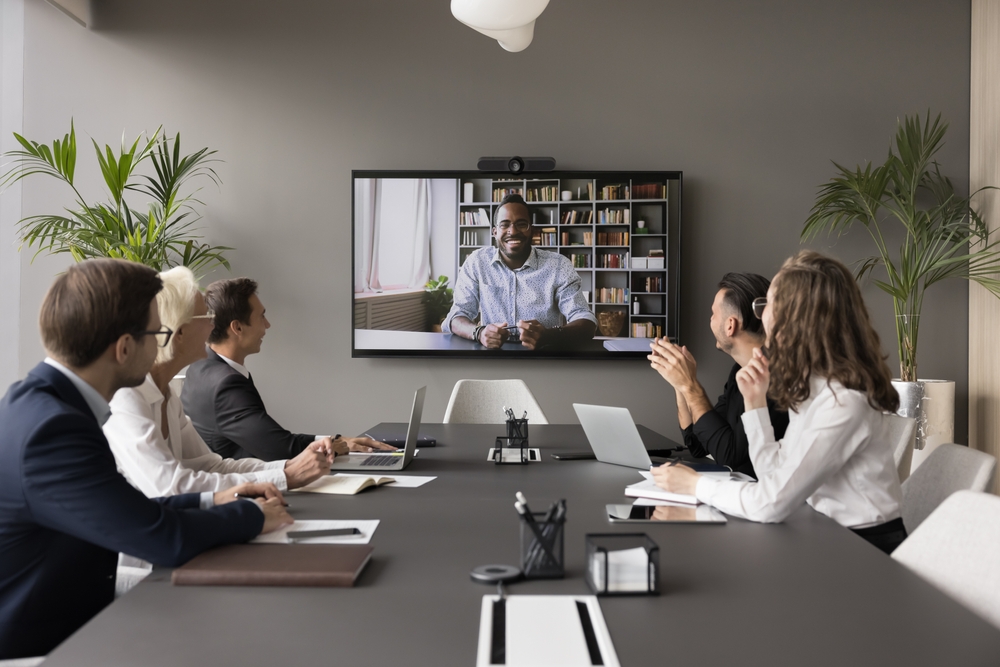 People in a conference room watch a man on a wall-mounted video screen during a meeting.