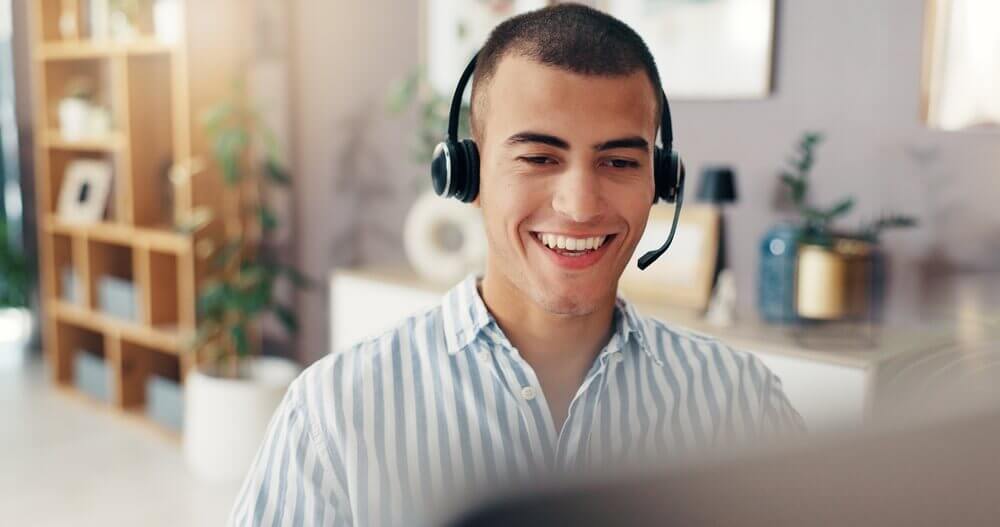 Smiling man wearing a headset and striped shirt at a computer in a bright home office.