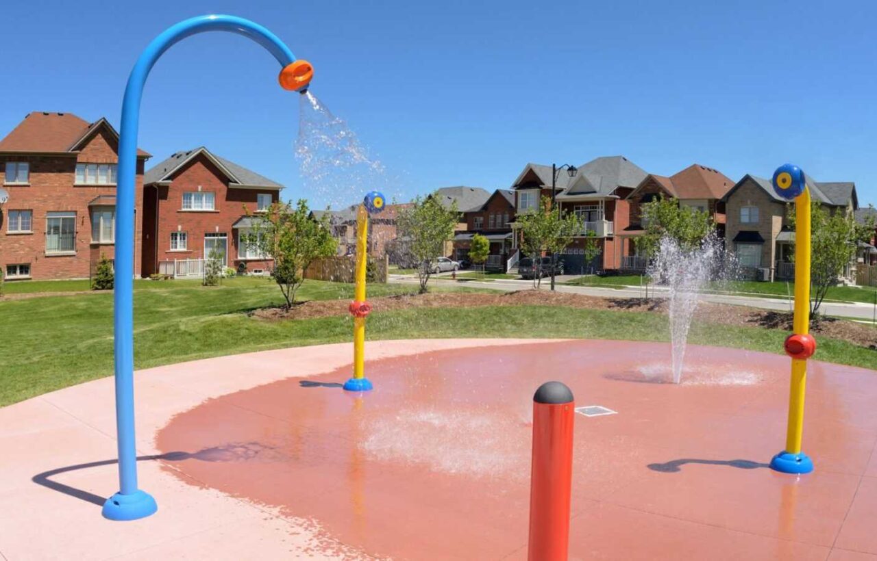 Outdoor splash pad with colorful water sprayers, wet red pavement, grass, and houses in the background