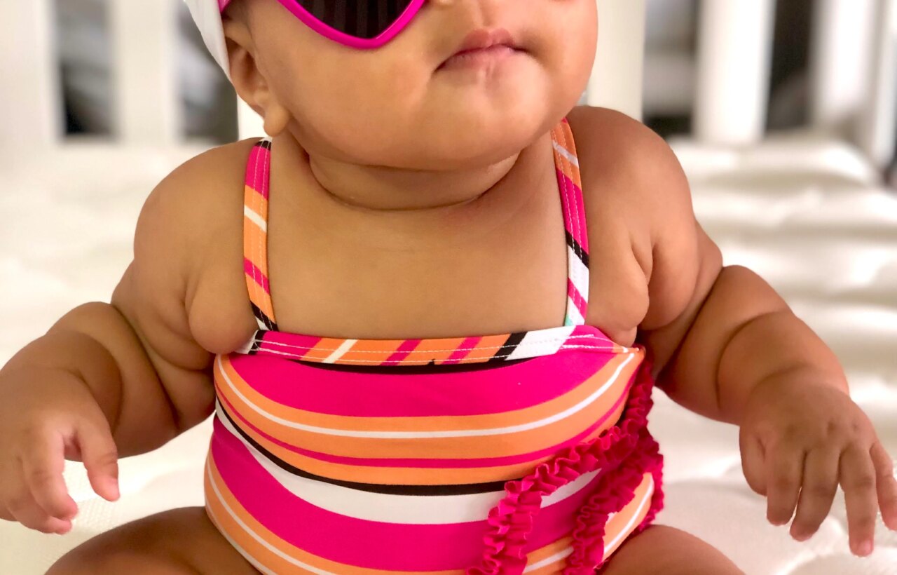 Baby wearing heart-shaped pink sunglasses, white headband, and striped swimsuit while sitting indoors