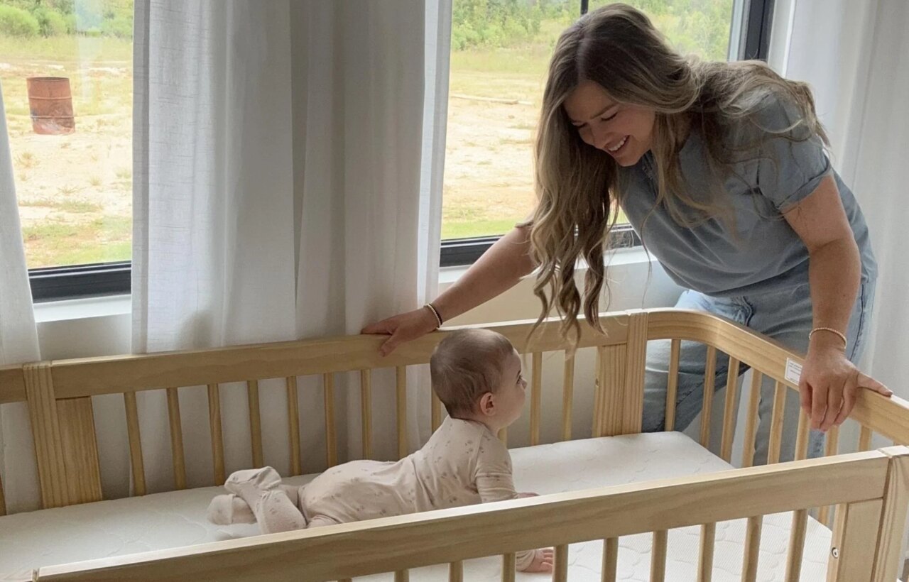 Woman leaning over a wooden crib toward a baby lying on a white mattress near large windows.