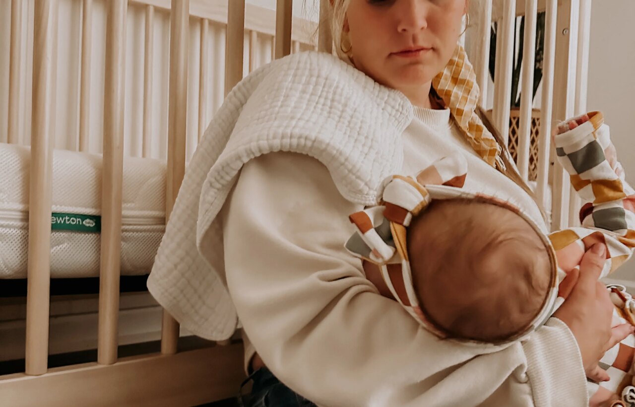 Adult holding a swaddled baby beside a wooden crib in a softly lit room