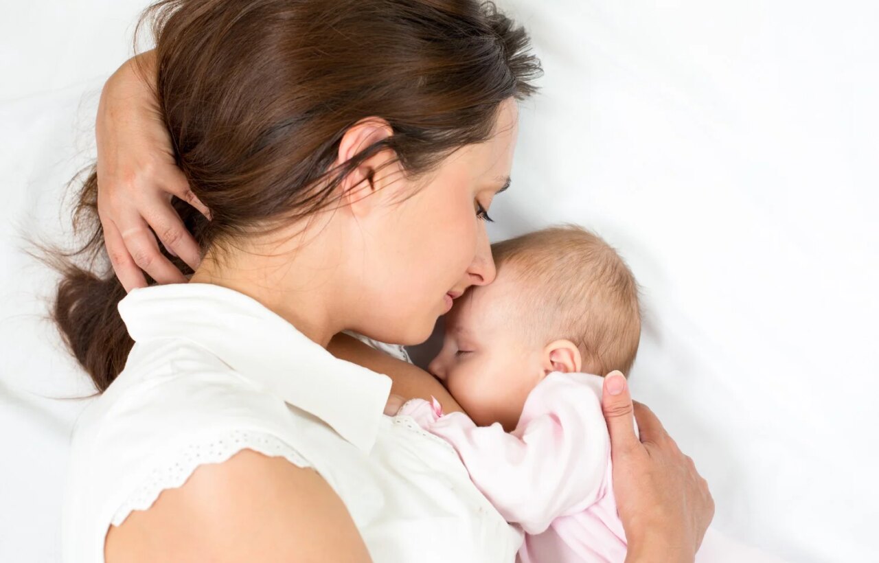Woman lying on white bedding holding a sleeping baby close to her chest.