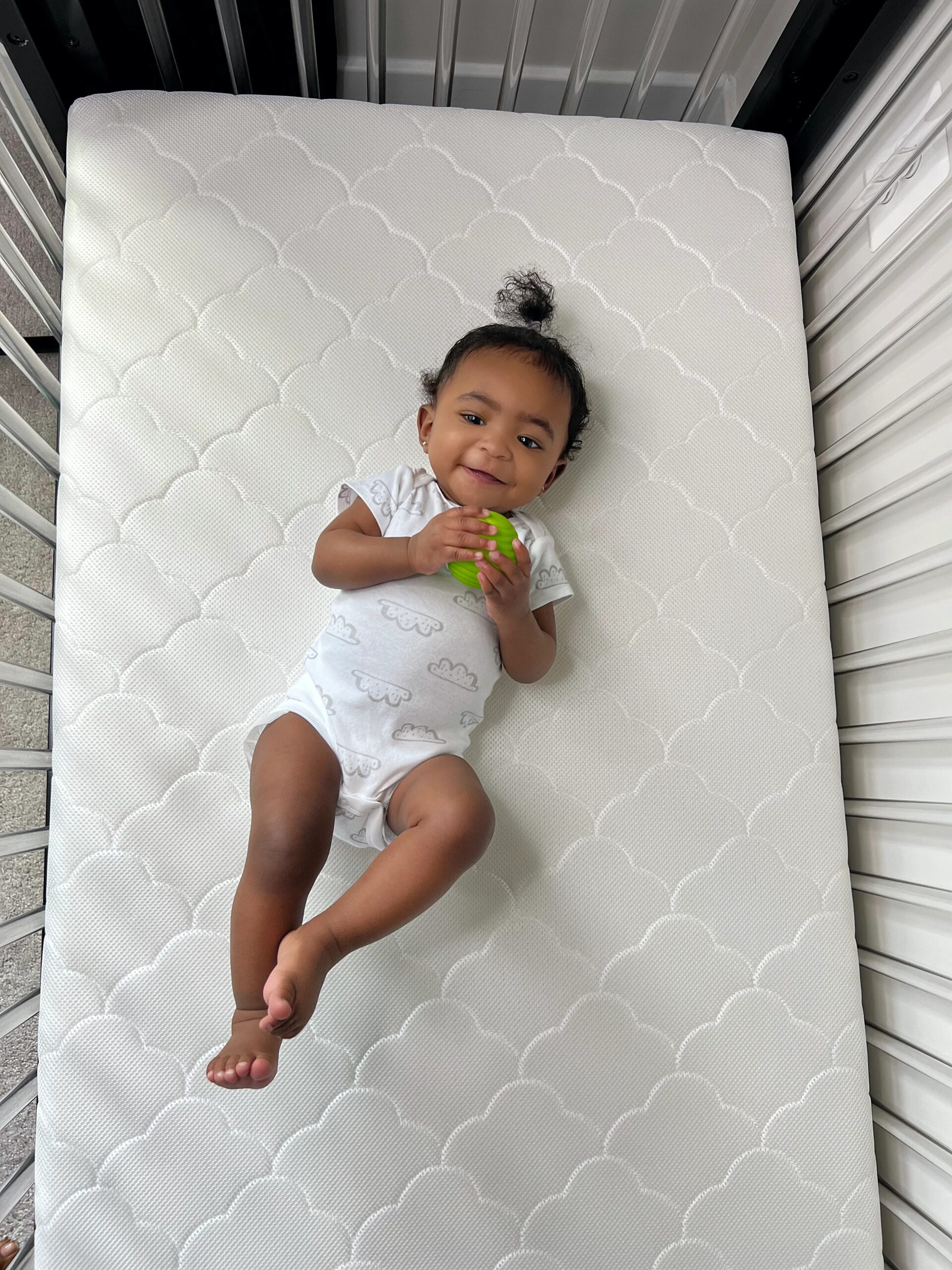 Smiling baby in a white onesie lying on a white crib mattress and holding a small green ball