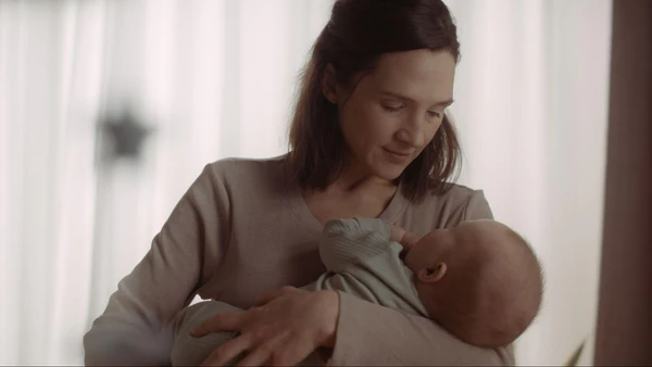 Woman looking down while holding an infant wrapped in a light blanket near bright curtains.