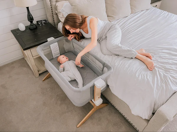 Woman on a bed reaching toward a baby in a bedside bassinet next to a nightstand.