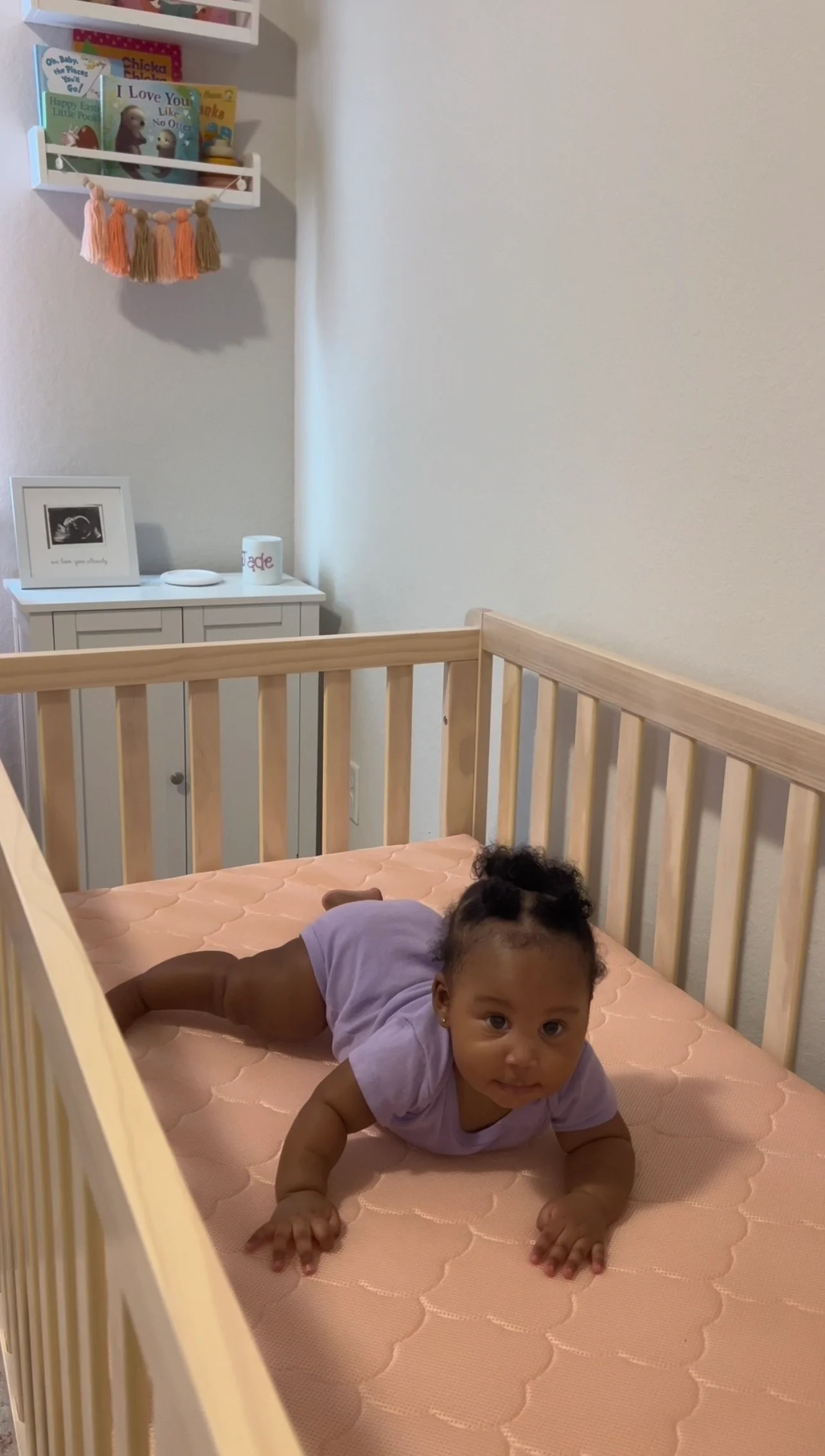 Baby lying on a pink crib mattress in a nursery with wall shelves, books, and a small cabinet.