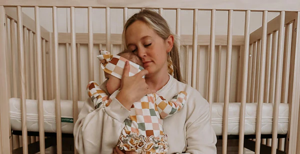 Woman holding a baby beside a wooden crib in a softly lit nursery