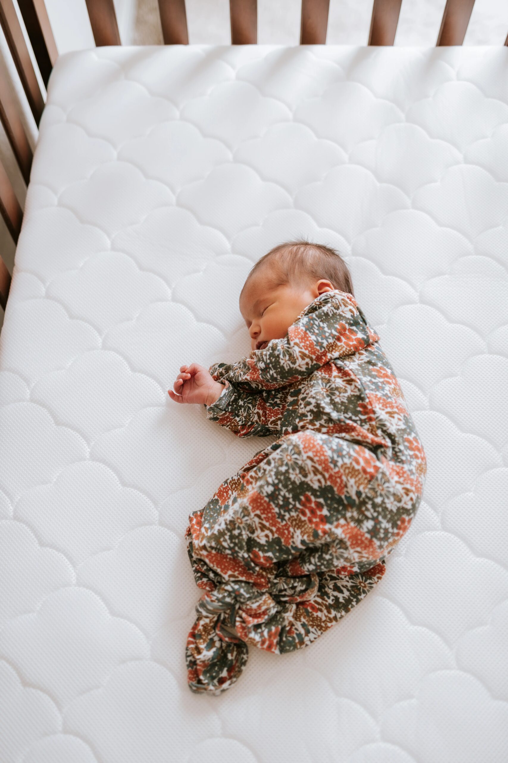 Sleeping baby in a floral outfit lying on a white quilted crib mattress.