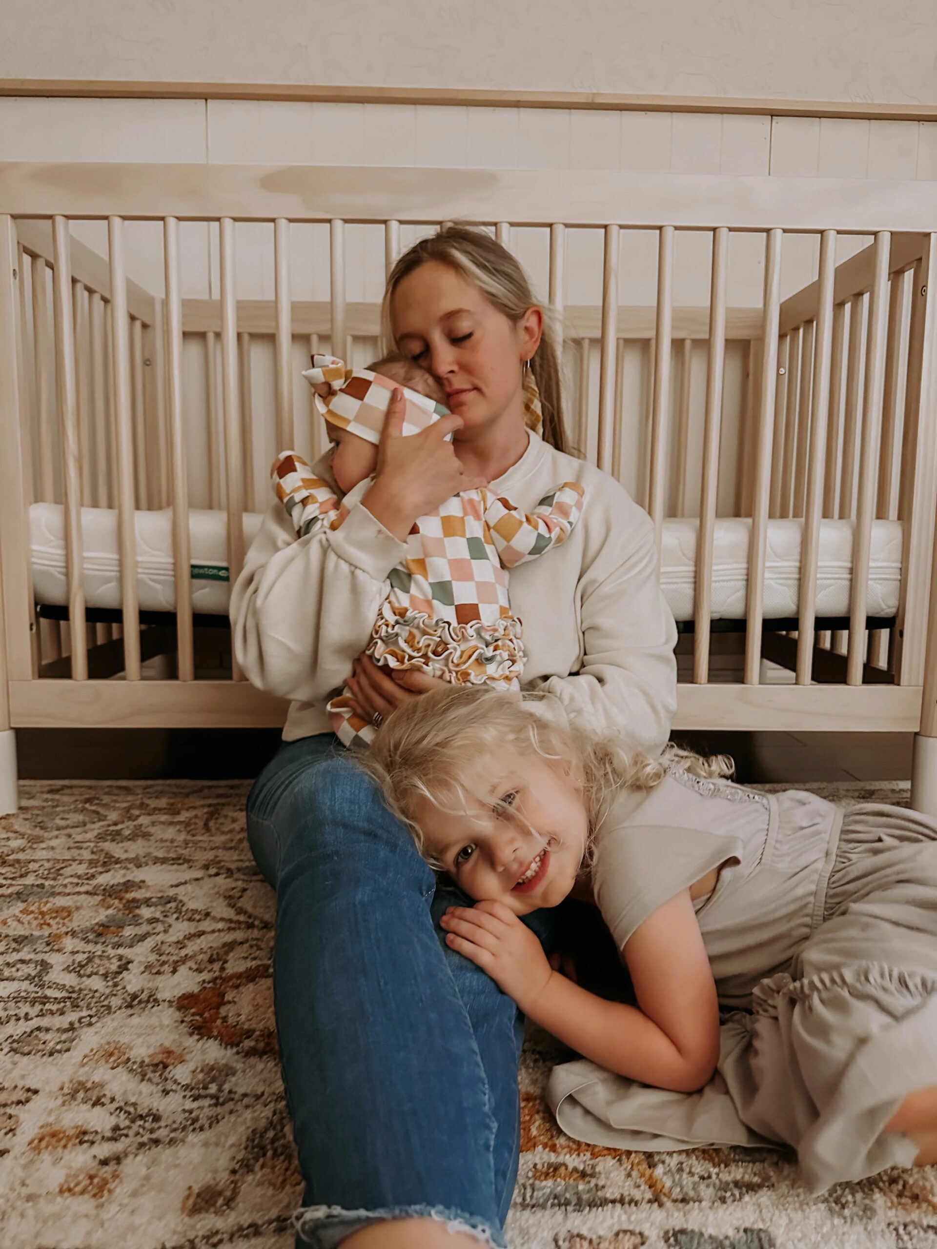 Woman holding a baby beside a crib while a young child rests her head on the woman's lap