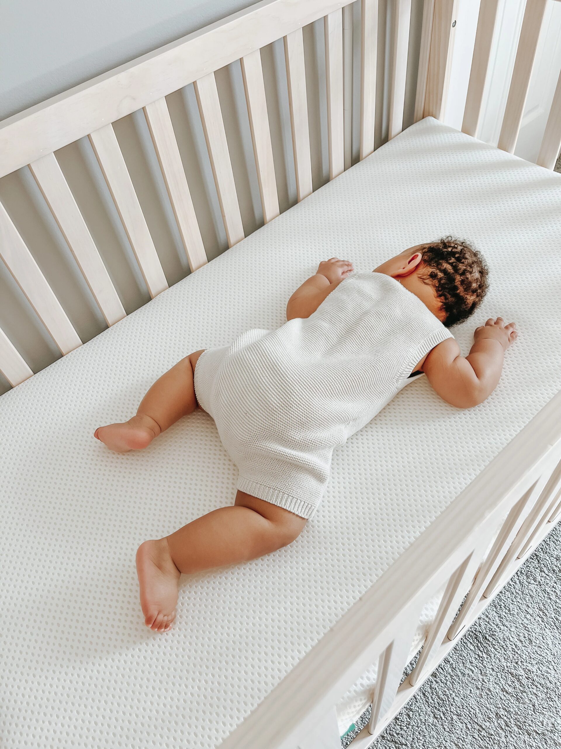 Baby lying on their stomach in a light wood crib on a white textured mattress.