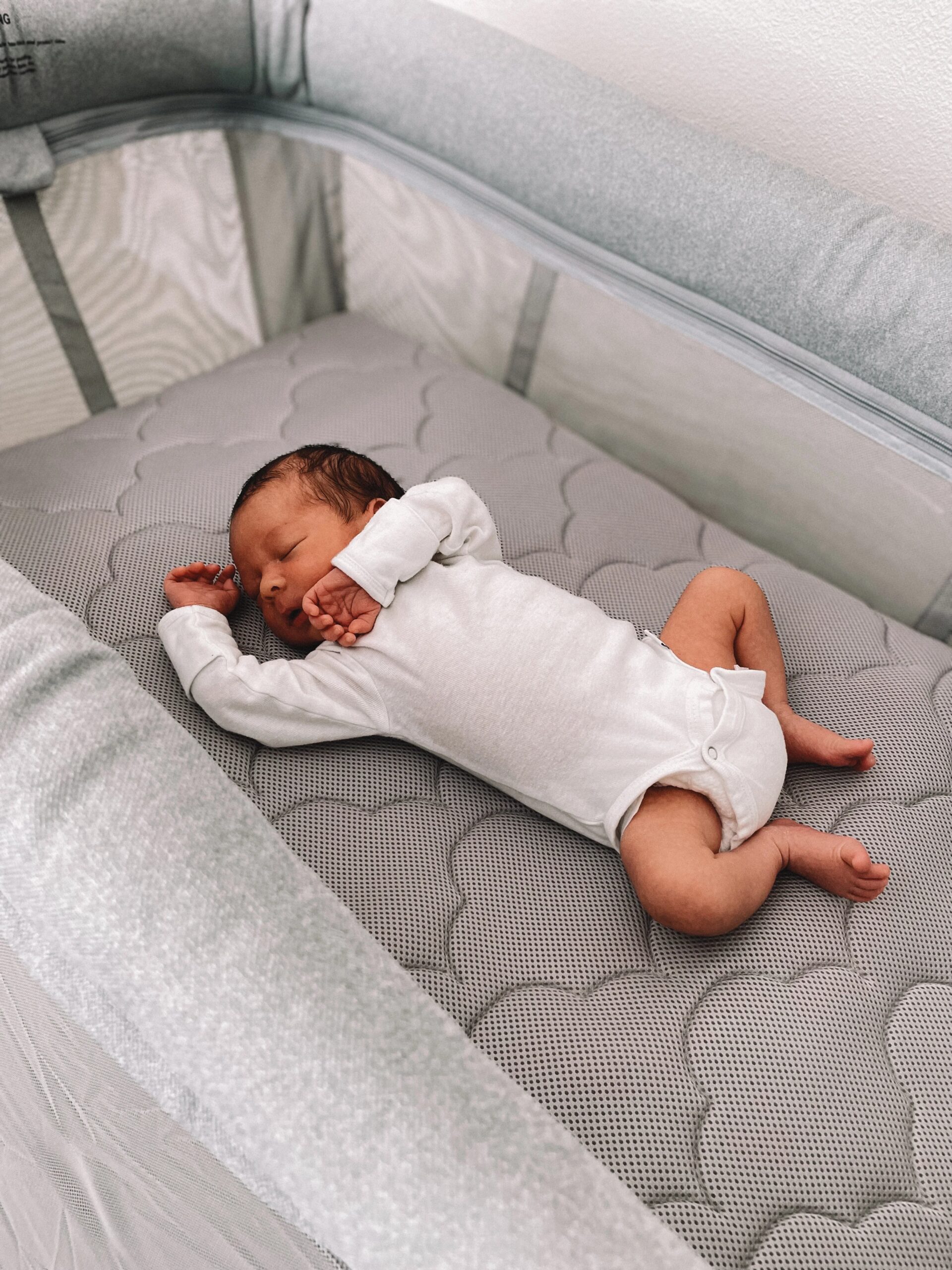 Sleeping infant in a white bodysuit lying on a gray quilted bassinet mattress.
