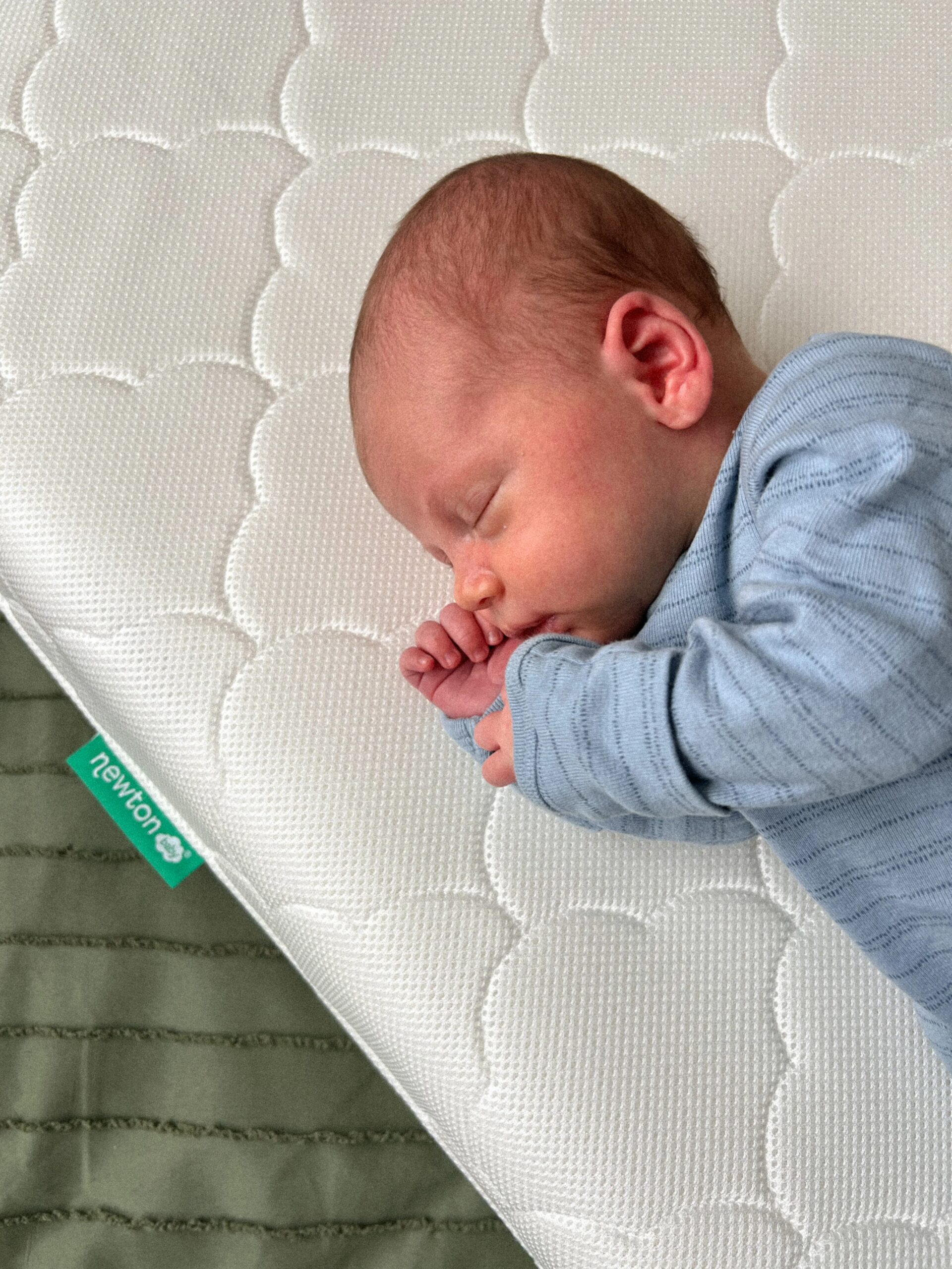 Sleeping baby in a blue outfit on a white quilted sleep surface with a Newton label