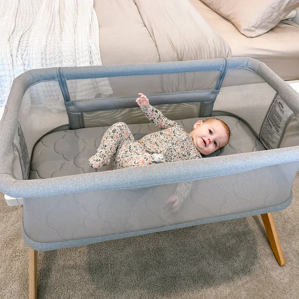 Smiling baby lying awake in a gray mesh bedside bassinet next to a bed