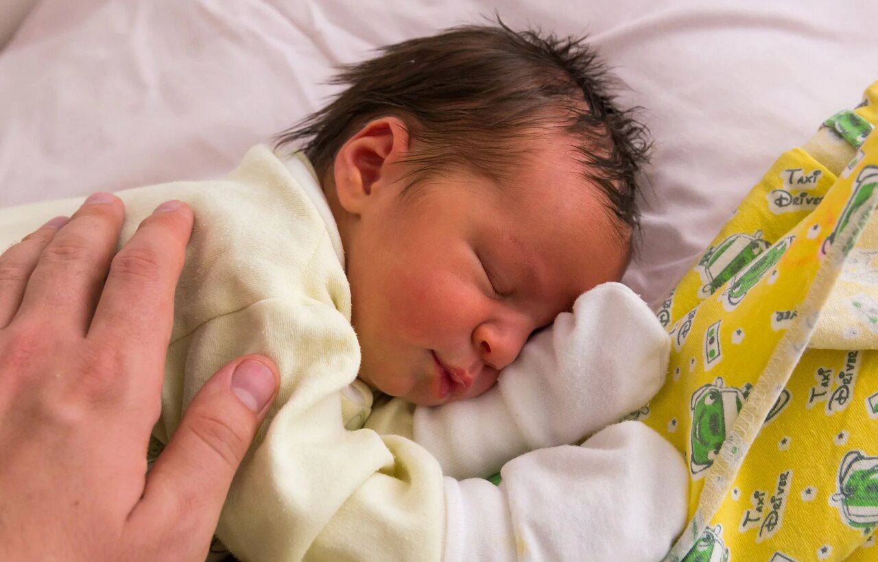 Sleeping newborn in pale clothing with an adult hand resting on their back