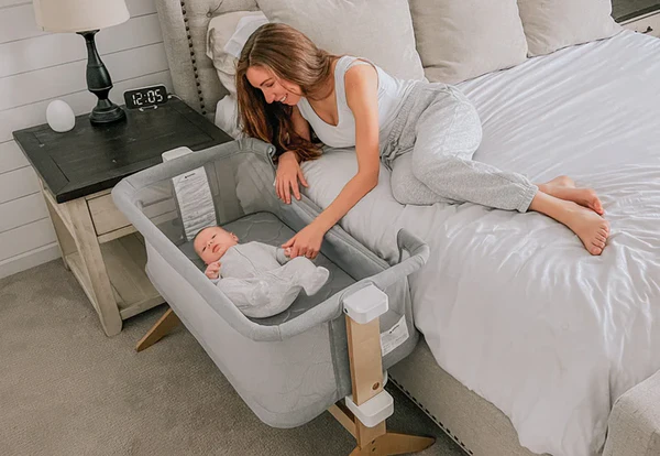 Woman lying beside a bed reaches toward a swaddled baby in a bedside bassinet.