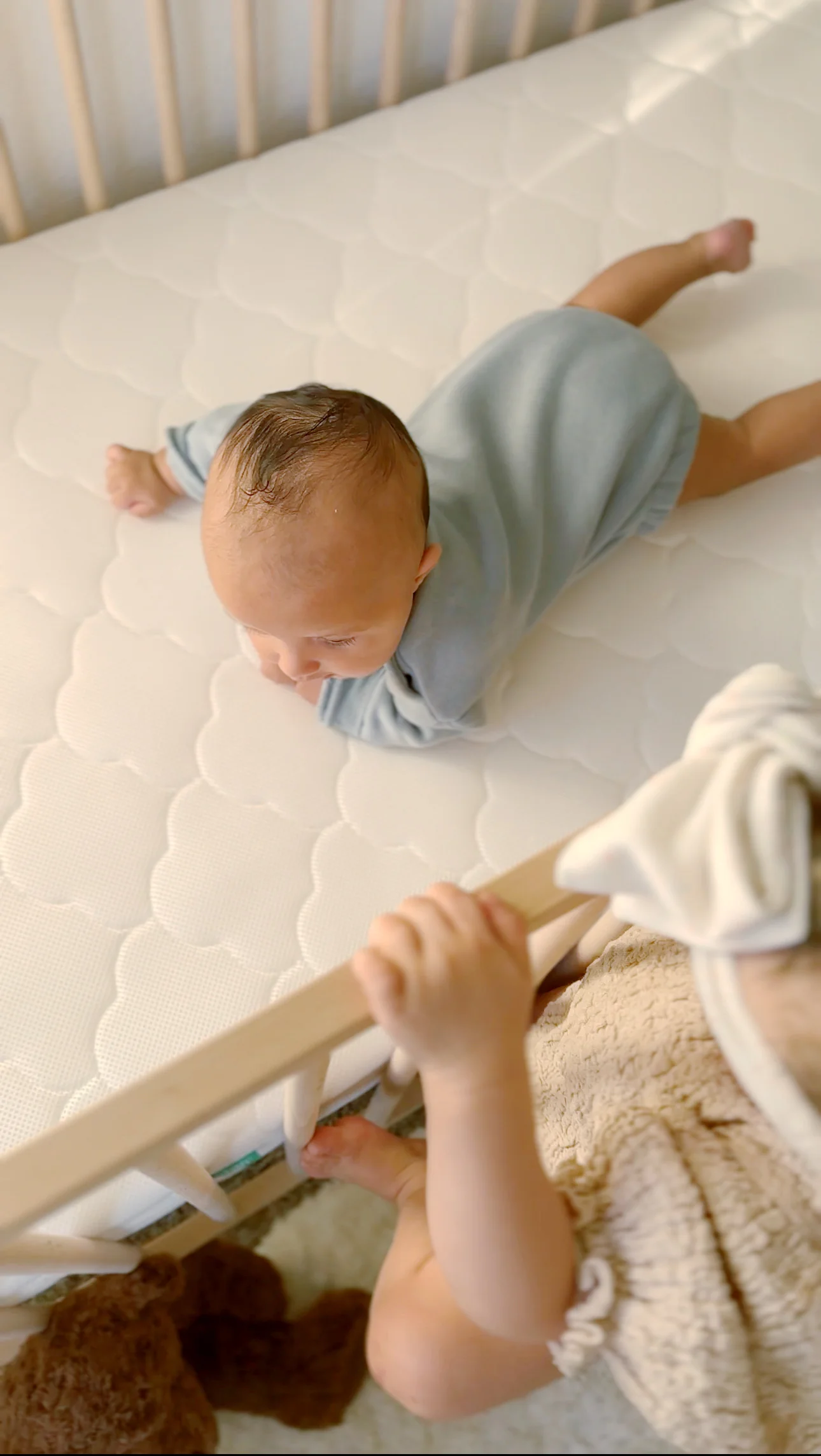 Infant lying on a white crib mattress while another child holds the crib rail nearby.