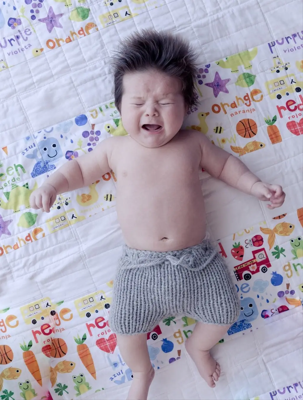 Crying baby lying on a colorful quilt, wearing gray knit shorts with eyes closed and hair standing up.