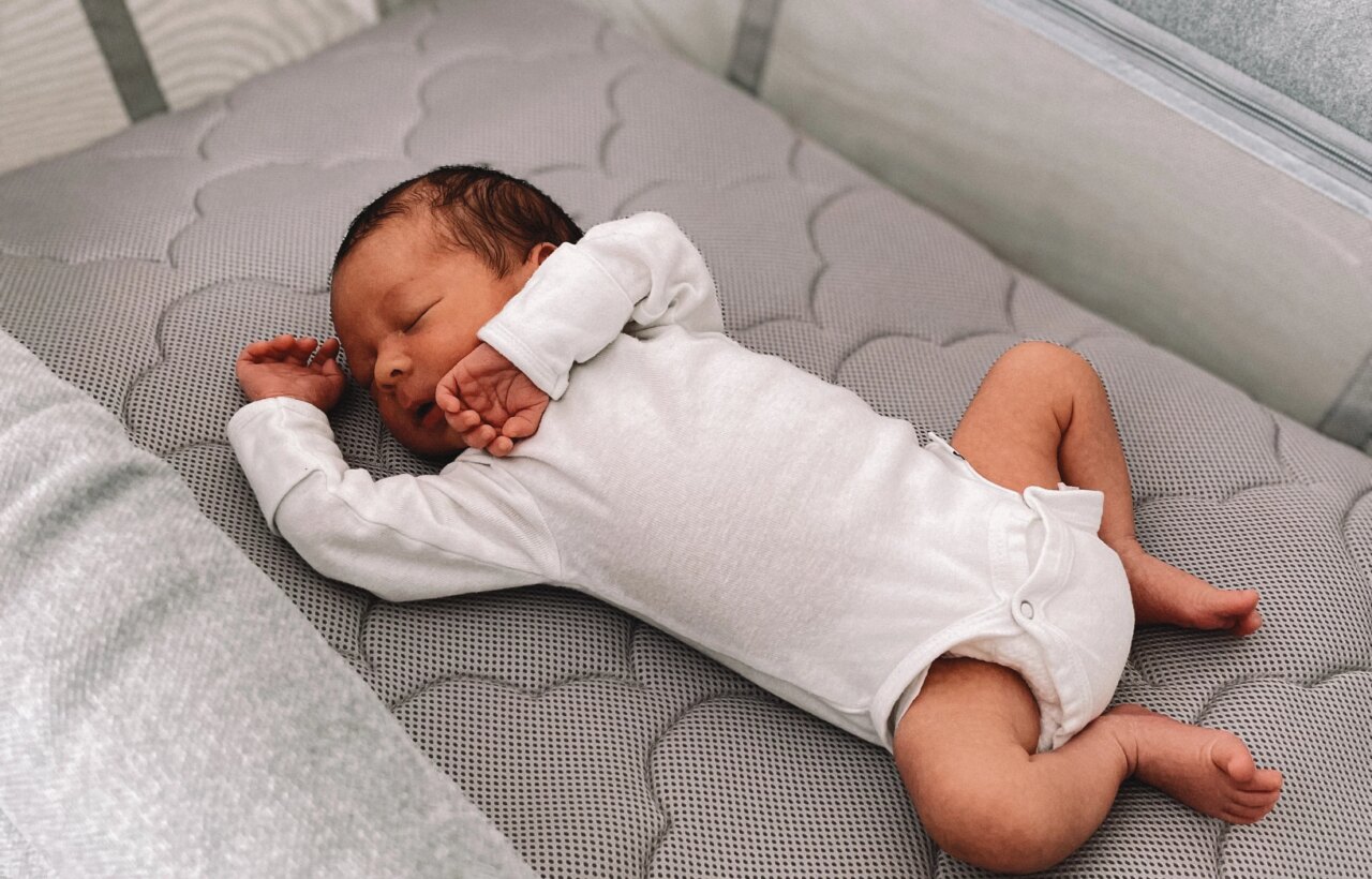 Sleeping infant in a white bodysuit lying on a gray quilted mattress inside a mesh-sided crib