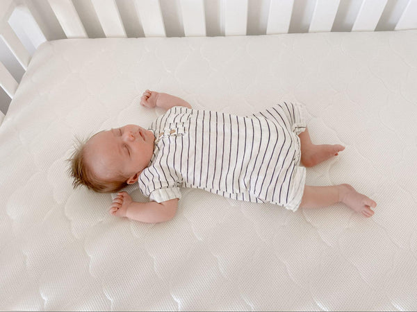 Baby lying on their back on a white crib mattress, wearing a striped sleep outfit.