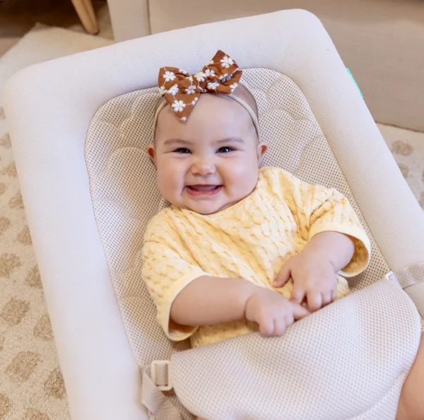 Smiling baby in a cushioned seat wearing a yellow outfit and brown floral bow headband.