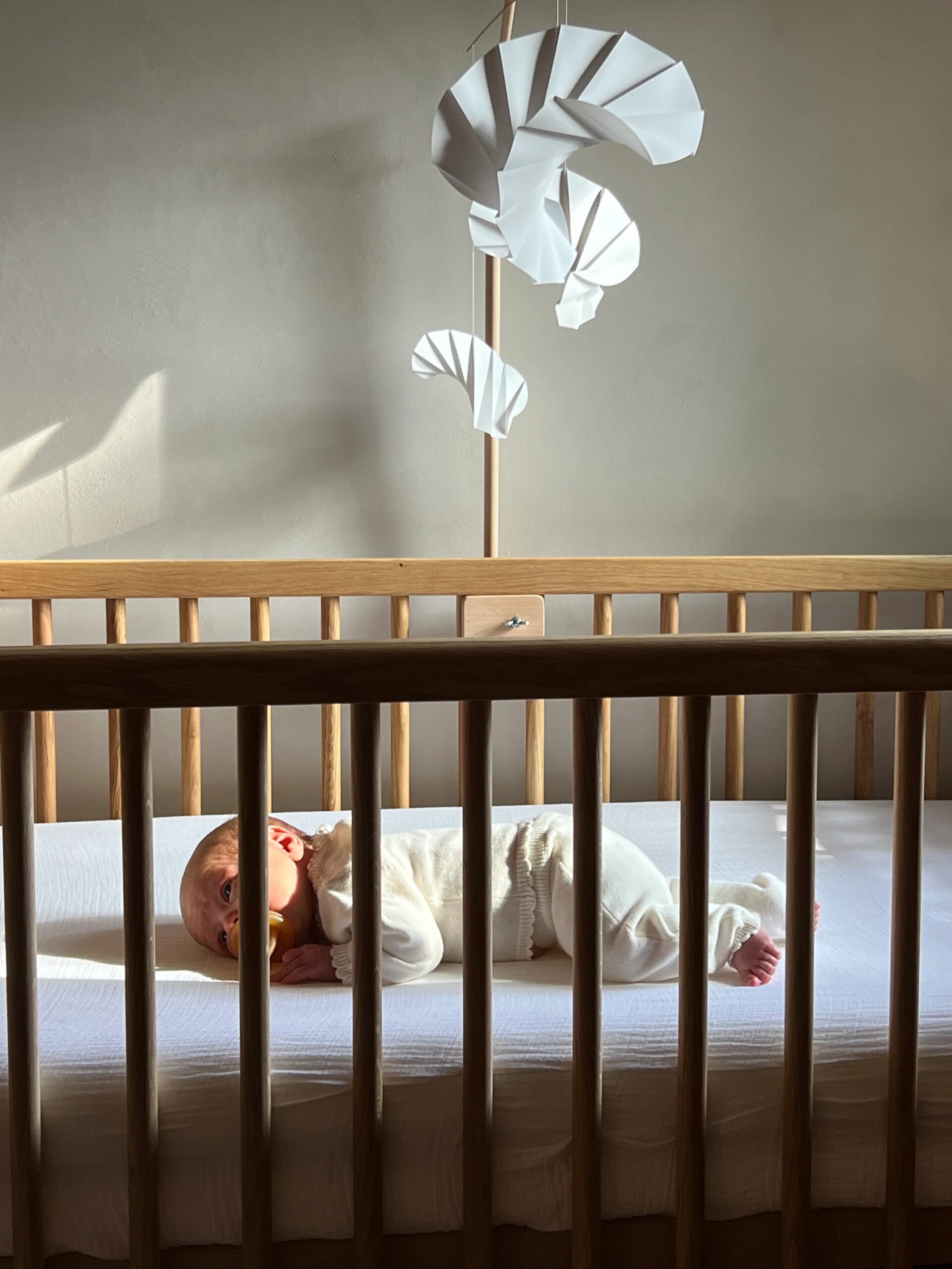 Baby lying in a wooden crib beneath a white hanging mobile in warm sunlight