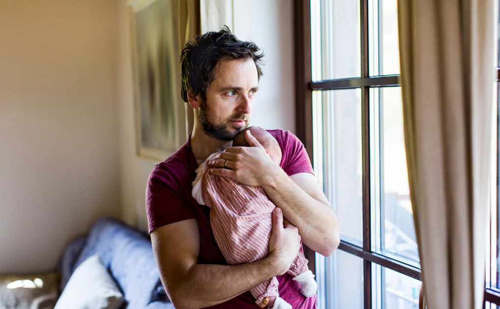 Man holding a baby against his chest while standing beside a window in a softly lit room.