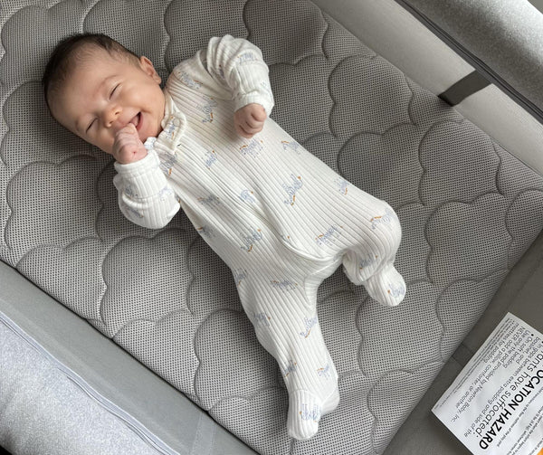 Baby with eyes closed lying on a gray quilted mattress in a white footed sleeper.