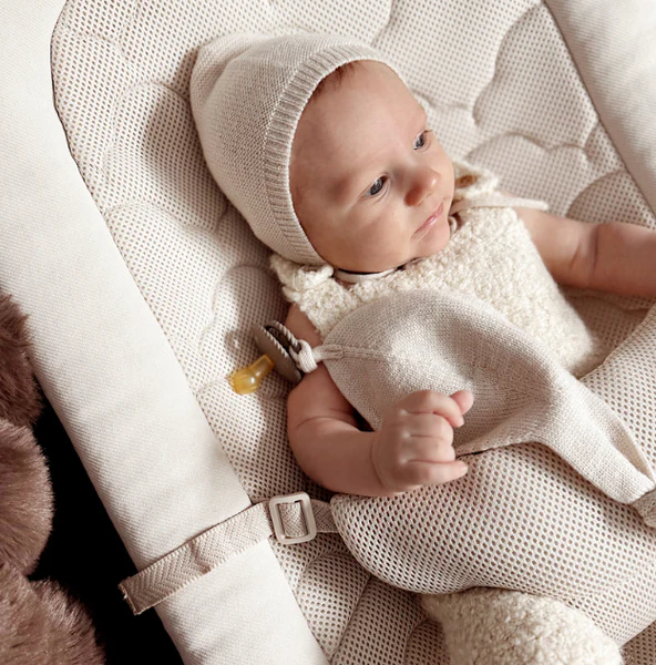 Baby in a beige mesh rocker wearing a knit bonnet and soft textured clothing