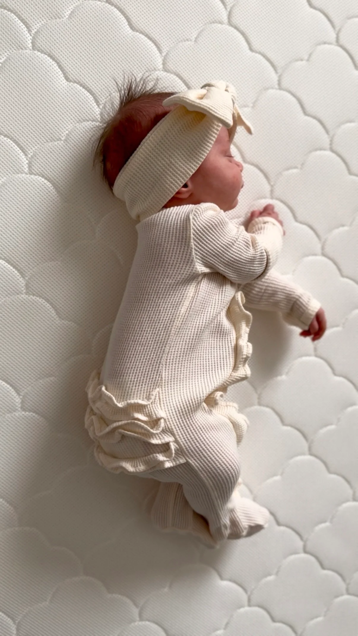 Newborn lying on their side on a white quilted mattress in a cream outfit and headband