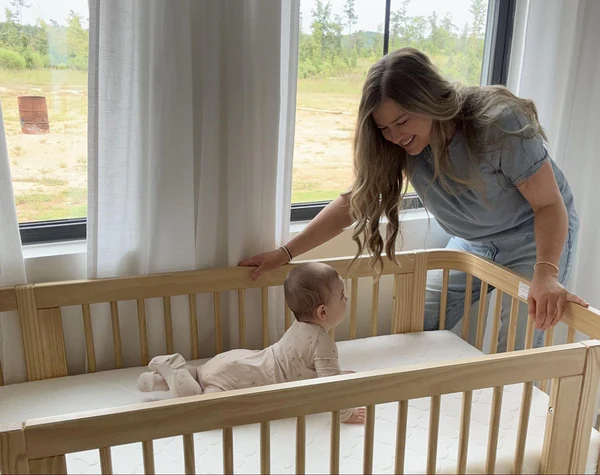 Baby lying on their stomach in a wooden crib while a smiling woman leans over the rail.