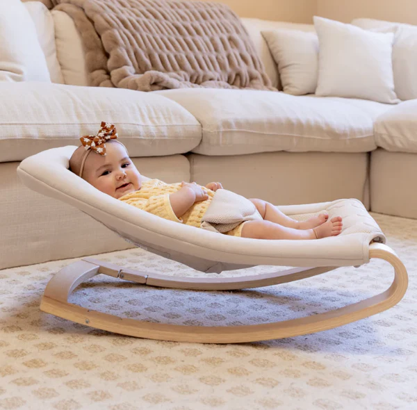 Baby lying in a cream rocker on a rug in front of a sofa
