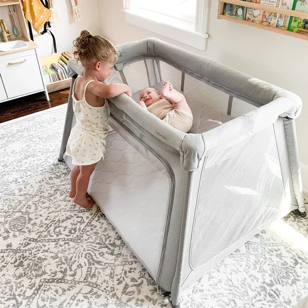 Toddler standing beside a gray mesh crib while a baby lies on their back inside.