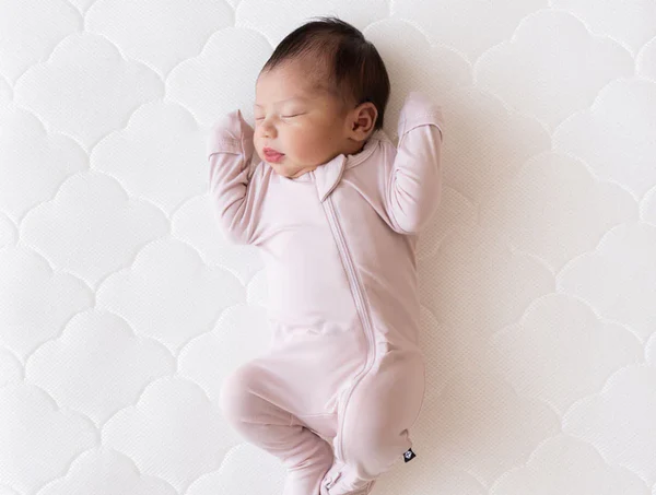 Sleeping newborn in a pale pink zip-up onesie lying on a white quilted mattress
