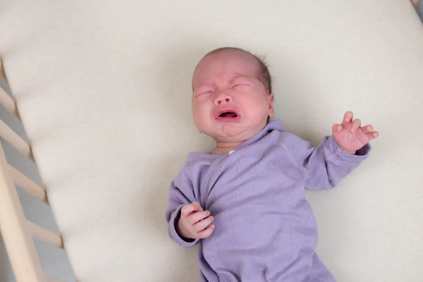 Crying baby in a purple outfit lying on a pale crib mattress