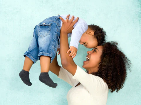 Smiling woman lifting a baby in denim overalls against a light teal background.