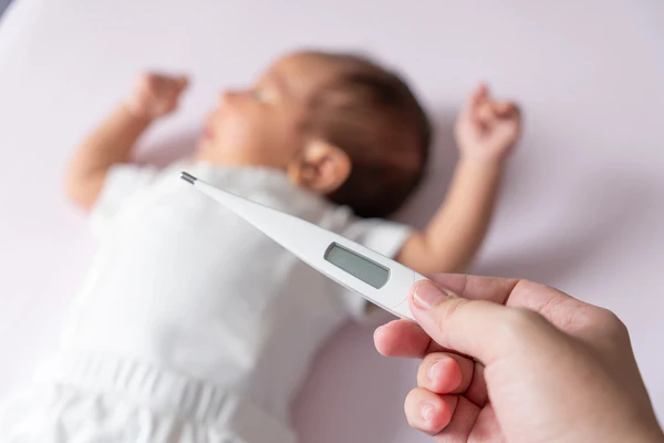 Hand holding a digital thermometer near a baby lying in bed