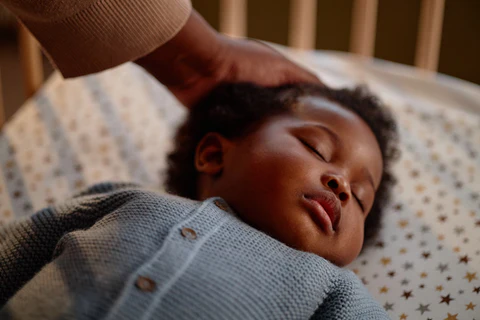 Sleeping baby in a blue buttoned outfit lying on a patterned sheet as an adult hand touches their head