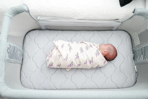 Swaddled infant lying on a gray quilted mattress inside a light gray bassinet.