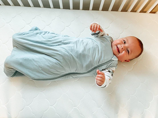 Smiling infant in a light blue swaddle lying on a white crib mattress.