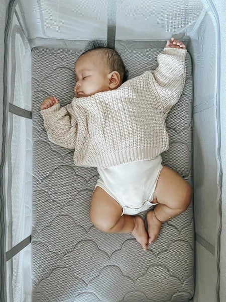Sleeping baby in a cream knit sweater lying on a gray patterned mattress in a mesh-sided bassinet.