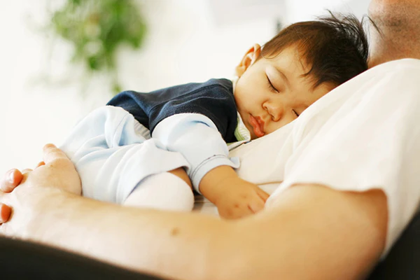 Sleeping baby resting on an adult's chest, with the adult's arm visible in a softly lit room