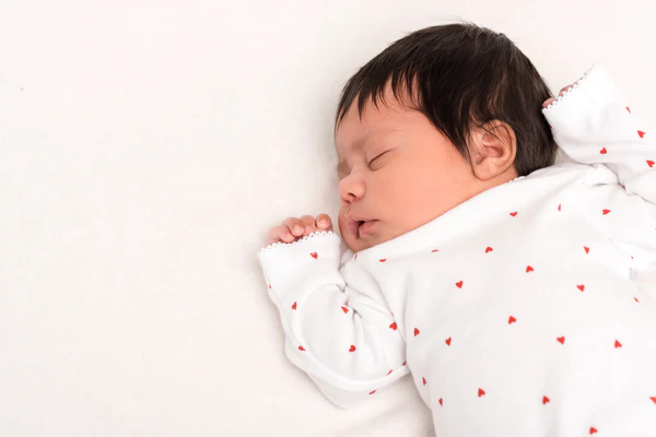 Sleeping baby in a white outfit with small red hearts, lying on white bedding