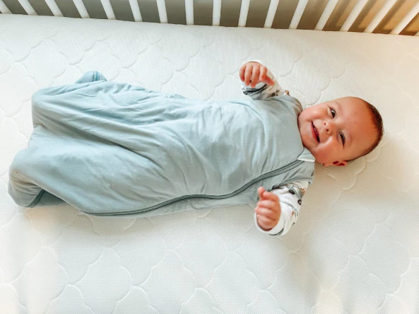 Smiling baby in a pale blue sleep sack lying on a white crib mattress