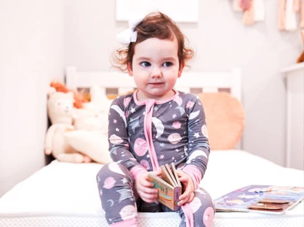 Young child in patterned pajamas sitting on a white bed holding a small board book