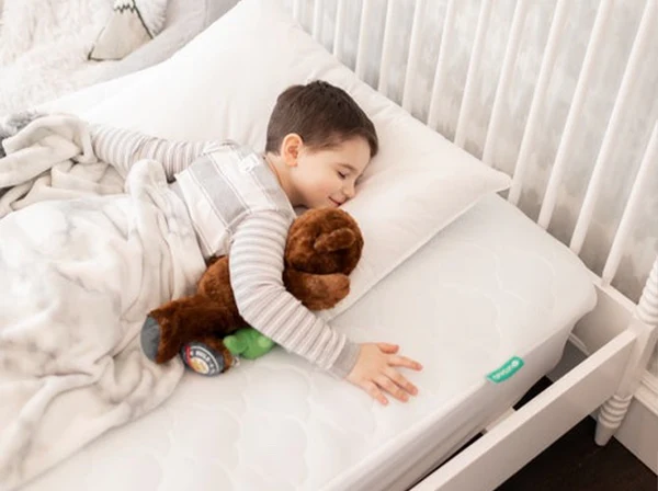 Young child asleep in a crib, hugging a brown stuffed animal on a white mattress.