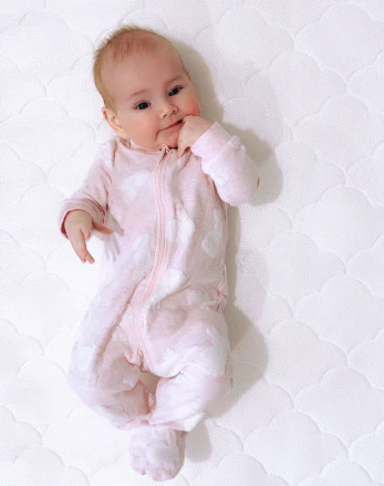 Baby in a pale pink sleeper lying on a white patterned mattress